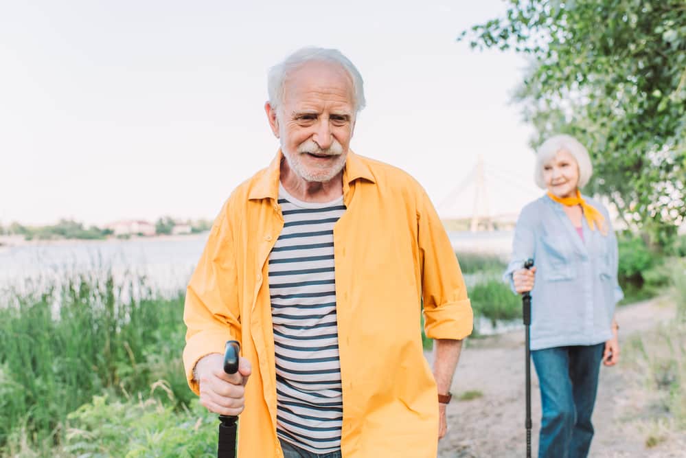 Senior-man-walking-outdoors-with-walking-stick-senior-woman-in-background