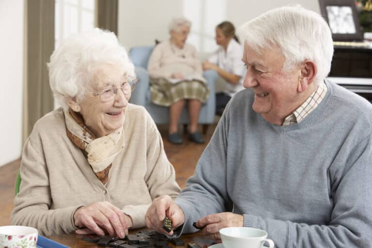 Senior couple smiling while playing dominos, senior woman and nurse in background